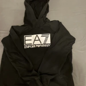 Svart EA7 Emporio Armani hoodie - Svart hoodie från EA7 Emporio Armani med stor vit logga framtill. Klassisk modell med huva och långa ärmar. Perfekt för dig som gillar streetwear och vill ha en stilren look med tydlig branding.