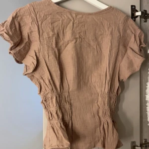 Beige blus med rynkade detaljer - Säljer en beige blus i mjukt bomullstyg med korta ärmar och rynkade partier i midjan som ger en snygg siluett. Blusen har en avslappnad passform och lätt struktur i tyget, perfekt för vår och sommar.