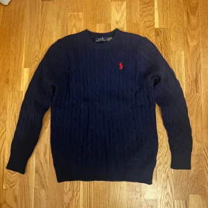 Mörkblå stickad tröja från Ralph Lauren - Kabelstickad mörkblå tröja från Ralph Lauren med klassisk röd broderad logga på bröstet. Tröjan har rund halsringning och långa ärmar. Perfekt för dig som gillar en tidlös och stilren look.