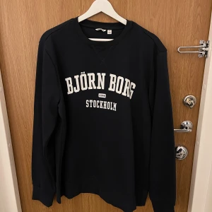 Svart sweatshirt från Björn Borg - Svart sweatshirt från Björn Borg med vit text på bröstet. Klassisk rund halsringning och långa ärmar. Perfekt för en avslappnad och sportig stil. Mjuk och bekväm, tillverkad i bomullsmix. Liten i storleken, passar M/L.