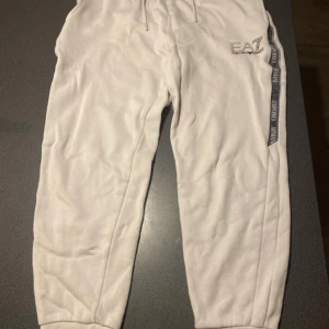 Beigea  mjukisbyxor EA7 Emporio Armani - Snygga vita mjukisbyxor från EA7 Emporio Armani med logga på benet och breda svarta sidoremsor med text. Byxorna har dragsko i midjan och muddar vid bensluten. Perfekta för en chill och sportig look ny pris 800kr.