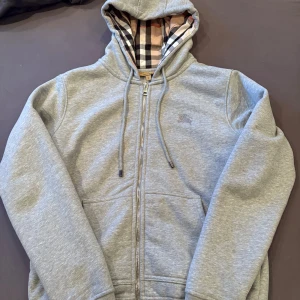 Burberry zipup  - Grå hoodie från Burberry med klassiskt rutigt foder i huvan, broderad logga på bröstet och hel dragkedja framtill. Tröjan har snörning i huvan och känguruficka. Perfekt för dig som gillar stilren design med lyxig touch. Använd fåtal gånger men är i absolut ny skick. Om du är minsta intresserad TVEKA INTE en sekund på att höra av dig, pris är absolut inte hugget i sten 