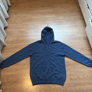 Mörkblå hoodie i kashmir - Mysig mörkblå hoodie i mjuk kashmir med långa ärmar och huva. Tröjan har ribbade muddar och dragsko i huvan för en avslappnad look. Perfekt för dig som gillar stilrena och bekväma plagg.