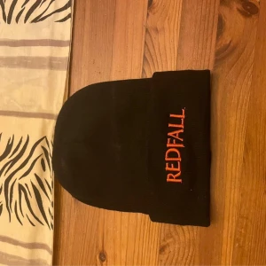 Svart Redfall mössa med brodyr - Svart mössa med uppvikt kant och broderad Redfall-logga i rött framtill. Klassisk, enkel design som passar till det mesta. Tillverkad i mjukt syntetmaterial och one size.