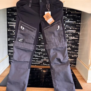 Svarta cargopants från RVRC - Grymma svarta cargopants från RVRC med gråa detaljer. Byxorna har flera dragkedjeförsedda fickor på benen och förstärkta partier. Perfekta för dig som gillar funktionella och snygga byxor med streetkänsla. Materialet är slitstarkt och passar för uteliv.