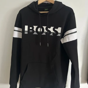 Svart hoodie från Hugo Boss med tryck - Svart hoodie från Hugo Boss med stort vitt BOSS-tryck på bröstet. Tröjan har huva med snörning och en stor magficka. 