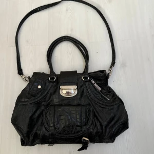 Vintage väska - Snygg svart vintage väska i croco-mönstrat skinnimitation. Väskan har både handtag och lång axelrem, silverfärgade metalldetaljer, flera fack med dragkedja och en cool frontdetalj. Perfekt för dig som vill ha plats för allt och ändå se stylish ut. Den är min mammas gamla så i andvänt men fint skick