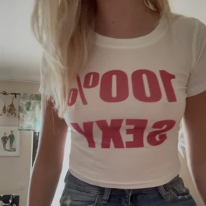 Vit croppad t-shirt med tryck - Säljer en vit croppad t-shirt med rosa texttryck '100% SEXY' på framsidan. T-shirten har rund hals och korta ärmar, sitter tight och är perfekt för dig som gillar att sticka ut. Materialet känns mjukt och stretchigt, troligtvis bomull eller polyester.