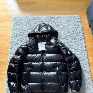 Moncler maya - Storlek 4. Snygg svart pufferjacka från Moncler med glansig finish och huva. Jackan har dragkedja framtill, två fickor med dragkedja och en ärmficka med Moncler-logga. Perfekt för kalla dagar och riktigt street. Klassisk Moncler-tag kvar. 