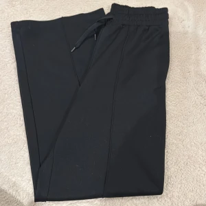 Svarta mjukisbyxor H&M Move 158/164 - Svarta mjukisbyxor från H&M Move med elastisk midja och dragsko. Byxorna har raka ben och sydda pressveck framtill för en clean look. Perfekta för chill dagar eller träning. Materialet är mjukt och stretchigt för extra komfort.