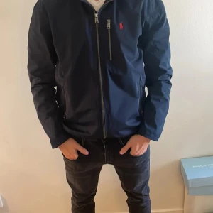 Ralph lauren vinjacka - Snygg mörkblå vindjacka från Polo Ralph Lauren med dragkedja framtill, huva och klassisk röd logga på bröstet. Jackan har flera fickor med dragkedja och en stilren, sportig look som passar perfekt till streetwear. Lätt och smidig att bära och är i storlek s