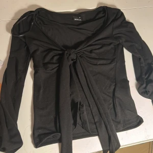 Svart långärmad topp med knyt Gina Tricot - Svart långärmad topp från Gina Tricot i storlek XS. Toppen har en snygg knytning framtill som ger en cool och trendig look. Mjuk och stretchig bomullskänsla, perfekt till jeans eller kjol. Djupare ringning och figurnära passform.
