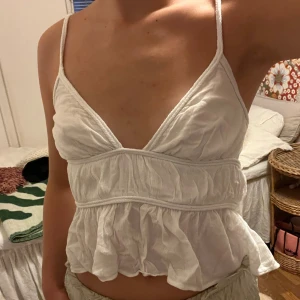 Vit volangig linnetopp med öppen rygg - Supersöt vit linnetopp med tunna axelband, volangig nederdel och v-ringning. Toppen har öppen rygg med knytband och är tillverkad i ett lätt, skrynkligt bomullstyg. Perfekt för varma dagar och ger en luftig känsla.