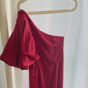 Röd oneshoulder långklänning Boohoo - Snygg röd långklänning från Boohoo med en dramatisk puffärm och oneshoulder-design. Klänningen har en elegant och stilren look, perfekt för dig som vill sticka ut. Tillverkad i ett mjukt, glansigt polyestertyg som faller snyggt.