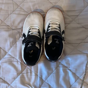 Vita och svarta sneakers från Hummel - Snygga sneakers från Hummel i vitt och svart med klassiska pilar på hälen. Ovandelen är i mesh och syntet, med snörning och kardborreband för extra support. Sulan är i gummi med bra grepp, perfekt för inomhussport eller vardag.