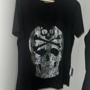 Svart Philipp Plein t-shirt med döskalle - Svart t-shirt från Philipp Plein med stor glittrig döskalle framtill, dekorerad med loggor och paljetter. Klassisk rund hals och korta ärmar. Snygg detalj med logotyp på ryggen och sidan. Perfekt för dig som gillar statement-plagg.