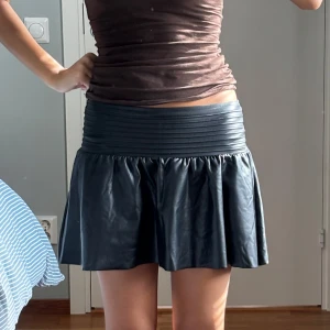 Svart kort kjol i skinnimitation Gina Tricot - Säljer en svart kort kjol från Gina Tricot i skinnimitation. Kjolen har en bred, ribbad midja och ett glansigt, lätt utställt tyg som ger en cool och edgy look. Passar XS 