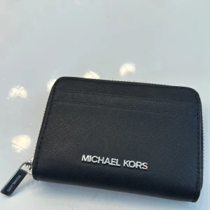 Svart plånbok från Michael Kors - Snygg svart plånbok från Michael Kors i stilren design med silverfärgad dragkedja och logga framtill. Plånboken har flera kortfack på utsidan och ett rymligt fack inuti. Tillverkad i tåligt skinnmaterial, perfekt för att hålla koll på dina kort och cash. I ny skick och endast använd några gånger, nypris 500kr säljer för 250kr.