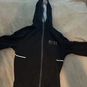 Svart hoodie från Hugo Boss med dragkedja - Svart hoodie från Hugo Boss med hel dragkedja framtill och vit logga på bröstet. Tröjan har huva med snörning och vita detaljer vid fickorna. Perfekt för en chill och stilren look.