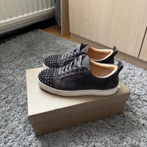 Christian Louboutin gråa  sneakers med nitar - Gråa  sneakers i mocka från Christian Louboutin med ikonisk röd sula och svarta nitar på tån. Skorna har snörning, rund tå och vit platt sula. Insidan är beige och materialet känns lyxigt. Perfekta för dig som vill sticka ut med en edgy vibe.