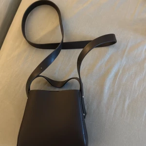 Svart axelväska i skinn med dragkedja - Stilren svart axelväska i slätt skinn med bred axelrem och minimalistisk design. Väskan har ett rymligt huvudfack och en innerficka med dragkedja för småsaker. Perfekt för dig som gillar enkel och modern look.