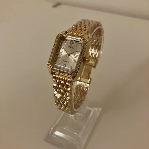 Guldig klocka från XIMILI med stenar - Snygg guldig klocka från XIMILI med fyrkantig urtavla och gnistrande stenar runt ramen. Länken är i metall och har en elegant design. Urtavlan är vit med romerska siffror och guldiga visare. Perfekt accessoar för att lyfta din stil.