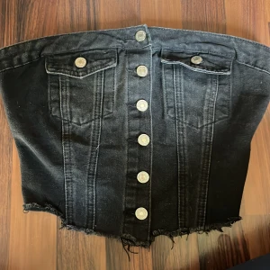 Svart jeans tubtopp från SHEIN - Cool svart tubtopp i jeansmaterial från SHEIN med knappar framtill, två fejkade bröstfickor och rå, fransig nederkant. Baksidan har smock för extra stretch och passform. Perfekt för dig som gillar edgy och trendiga plagg.