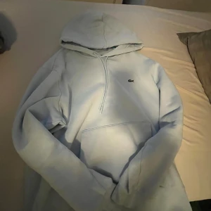 Ljusblå hoodie från Lacoste - Säljer en ljusblå hoodie från Lacoste med klassisk krokodillogga på bröstet. Tröjan har huva med snörning, stor magficka och långa ärmar. Perfekt för chill dagar och enkel att matcha med jeans eller joggers. Bäst att mötas upp