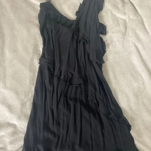 Svart omlottklänning med volanger H&M XS - Svart kortklänning från H&M i storlek XS. Klänningen har ett omlottliknande snitt med volangdetaljer och asymmetrisk design. Axelbandslös på ena sidan och tunna axelband på andra ger en cool oneshoulder-look. Mjuk och lätt polyester som faller snyggt.