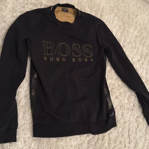 Svart sweatshirt från Hugo Boss - Svart sweatshirt från Hugo Boss med stort guldigt BOSS-tryck på bröstet och diskreta logodetaljer längs sidorna. Tröjan har rund hals, långa ärmar och ribbade muddar. Perfekt för dig som gillar stilrena och exklusiva plagg.