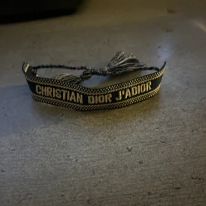 Christian Dior J'Adior armband - Snyggt armband från Christian Dior med texten 'J'Adior' broderad i vitt på svart tyg. Armbandet har justerbar knytning med tofsar i ändarna och en cool, bohemisk känsla. Perfekt accessoar för att lyfta din outfit med en lyxig touch.