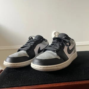 Nike Air Jordan 1 Low svart/grå - Säljer ett par Nike Air Jordan 1 Low i svart, grått och vitt skinn. Klassisk siluett med svart swoosh, broderad Air Jordan-logga på hälen och perforerad tå. Låga sneakers med snörning och platt sula – perfekta för dig som gillar streetstyle.