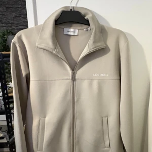 Beige ziptröja från Les Deux - Säljer en stilren beige ziptröja från Les Deux med hög krage och diskret logga på bröstet. Tröjan har hel dragkedja framtill, två fickor och är långärmad. Perfekt för lager-på-lager och enkel att matcha med det mesta.