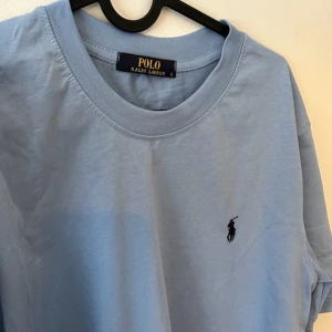 Ljusblå t-shirt från Polo Ralph Lauren - Snygg ljusblå t-shirt från Polo Ralph Lauren med klassisk rund hals och broderad logga på bröstet. T-shirten är i mjuk bomull och har en clean, stilren look som passar perfekt till jeans eller shorts.