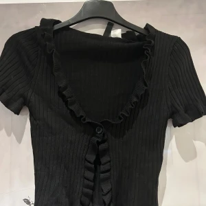 Svart ribbad topp med volang - Supersöt svart ribbad topp med volangdetaljer längs v-ringningen, ärmsluten och framtill. Toppen har en knapp framtill och är croppad i modellen. Storlek XS från hm!🖤