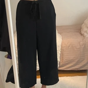 Svarta vida byxor med snörning - Svarta byxor med breda ben och snörning i midjan. De har en croppad längd och är tillverkade i ett mjukt, lätt material som ger en avslappnad och trendig look. Perfekta för dig som gillar en loose och bekväm stil.