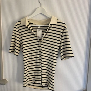 Randig ribbad t-shirt med krage - Snygg ribbad t-shirt från H&M i storlek M, med svart och vit randigt mönster. Modellen har korta ärmar, klassisk krage och knappar framtill. Perfekt för dig som gillar en clean och stilren look med lite preppy vibe.