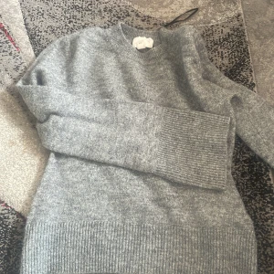 Grå stickad tröja från H&M - Mysig grå stickad tröja från H&M i storlek S. Tröjan har rund halsringning, långa ärmar och breda ribbade muddar vid ärmslut och nederkant. Perfekt för lager-på-lager och chill dagar.