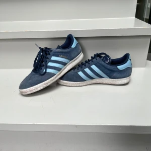 Adidas Gazelle blå sneakers i mocka - Säljer ett par Adidas Gazelle sneakers i mörkblå mocka med ljusblå detaljer och klassiska tre ränder på sidorna. Skorna har snörning, platt sula och rund tå. Adidas-logga bak i ljusblått och guldtext på sidan. Perfekta för dig som gillar retrostil och streetwear.