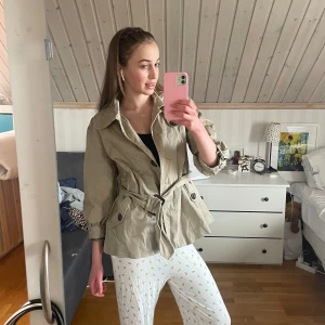 Beige trenchcoat från H&M med bälte - Snygg beige trenchcoat från H&M i storlek 40. Jackan har knappar framtill, bälte i midjan och trekvartsärmar. Insidan är fodrad med blommigt tyg i grönt och brunt. Perfekt för vår och höst när du vill ha en chill men stilren look.