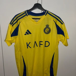 Al Nassr Ronaldo 7 matchtröja Adidas M - Gul och blå Al Nassr fotbollströja från Adidas med Ronaldo 7-tryck på ryggen. Slim fit-modell med korta ärmar, klubbmärke och sponsortryck framtill. Tillverkad i lätt och ventilerande polyester, perfekt för match eller träning.