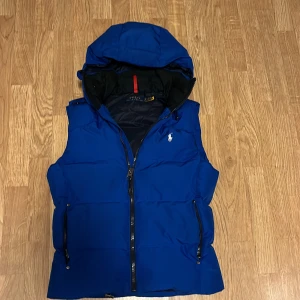 Blå dunväst från Polo Ralph Lauren - Snygg blå dunväst från Polo Ralph Lauren med huva och dragkedja framtill.Väldigt gott skick sparsamt använd då jag bara haft den i tre månader. Är i storleken XS men passar som en S