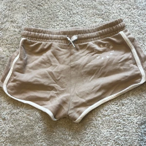 Beige shorts med vit kant från H&M - Säljer ett par beige shorts från H&M med vit kant och snörning i midjan. De har en sportig look och är tillverkade i mjuk bomull, perfekta för varma dagar. Passformen är normal och de har elastisk midja för extra komfort. Kontakta för fler bilder 🩷