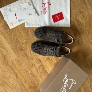 Christian Louboutin svarta sneakers mocka - Säljer ett par svarta sneakers från Christian Louboutin i mocka med ikoniska röda sulor. Skorna har snörning och coola nitar på tån för en edgy vibe. Insidan är beige och de har en platt sula. Perfekta för dig som vill sticka ut med lyxig streetstyle.