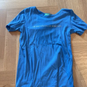 Blå t-shirt från United Colors of Benetton - Blå t-shirt med rund halsringning och korta ärmar från United Colors of Benetton. Tryckt logga i silver framtill. Skön och mjuk i bomull, perfekt för en avslappnad stil.
