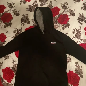 Svart hoodie från McKenzie XL - Svart hoodie från McKenzie med vit logga på bröstet. Klassisk modell med huva och långärmade ärmar. Insidan av huvan är grå. Perfekt för chill dagar eller när du vill ha en enkel och stilren look.