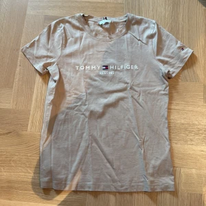 Beige t-shirt från Tommy Hilfiger - Snygg beige t-shirt från Tommy Hilfiger med klassisk logga och text framtill. T-shirten har rund halsringning och korta ärmar. Tillverkad i mjuk bomull som känns skön mot huden. Perfekt för en clean och stilren look.