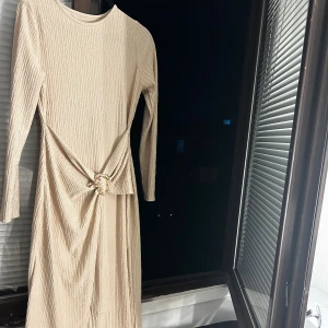 Beige långklänning med gulddetalj - Snygg beige långklänning med lång ärm och cool struktur i tyget. Klänningen har en unik guldfärgad detalj i midjan som ger en extra touch. Materialet känns lätt och har en skön textur. Perfekt för dig som vill sticka ut med stilren elegans.