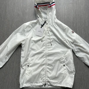 Vit vindjacka från Moncler med huva - Snygg vit vindjacka från Moncler med dragkedja, huva med snörning och coola röd-blå ränder på insidan av huvan. Jackan har två fickor med tryckknappar och Moncler-logga på ärmen. Perfekt för dig som vill ha en clean och stilren look.