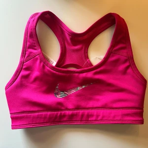 Nike sport-bh i rosa och mönster - Nike sport-bh med två sidor: ena helcerise med randigt Nike-logga, andra sidan har ett färgglatt randigt mönster i rosa, svart, grått och grönt. Tight passform, brottarrygg och tillverkad i stretchigt syntetmaterial. Perfekt för träning och sportiga aktiviteter. OBS aldrig använd! Råkade hamna i tvättmaskinen så prislappen åkte av.. 